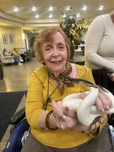 An elderly woman in a yellow shirt smiles while holding an exotic animal—a white snake—in her hands inside a well-lit room.