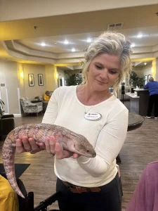 A woman wearing a name tag holds a large, pinkish exotic animal in a well-lit indoor setting, creating a safari-like atmosphere.
