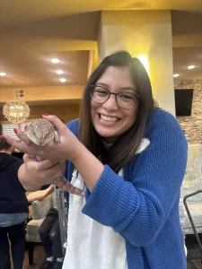 A woman wearing glasses and a blue cardigan smiles while holding an exotic animal lizard indoors, bringing a touch of exotic safari adventure to her day.