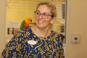 A woman wearing glasses and a floral blue dress smiles as an exotic animal—a colorful parrot—perches on her shoulder, evoking the spirit of a safari adventure.