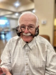 An elderly man with glasses smiles as two small, dark-colored bats cling to the sides of his face, capturing a unique moment reminiscent of an exotic animal safari.