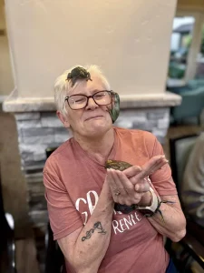 Older woman wearing glasses and a red shirt sits smiling with an exotic animal, a chameleon on her head, another on her face, and a frog resting on her hands—capturing the spirit of a personal animal safari.