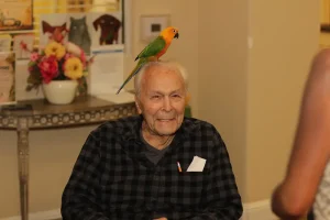 An elderly man sits and smiles with an exotic animal—a parrot—perched on his head; a table with flowers and framed pictures is in the background.