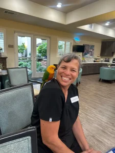 A smiling woman wearing black scrubs sits indoors with a colorful parrot perched on her shoulder, capturing the spirit of an Exotic Animal Safari; tables and chairs are visible in the background.