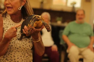A woman holds up a tortoise and points at it during an Exotic Animal Safari, while people sit in the background, watching attentively.