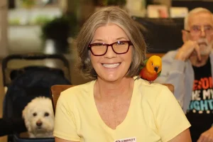 A woman with glasses smiles at the camera, a parrot perched on her shoulder as if she's on an Exotic Animal Safari. A white dog sits in the background, and an older man is also visible behind her.