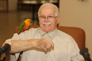 An elderly man with glasses and a mustache sits in a chair, smiling warmly, as if recalling tales from his Exotic Animal Safari, with a small colorful parrot perched on his arm.
