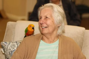 An elderly woman with white hair sits on a beige couch, smiling. An exotic animal—a colorful parrot—is perched on her shoulder, adding a touch of safari adventure to the cozy scene.