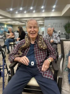 An elderly man sits in a chair with an exotic animal—a snake—draped around his shoulders, smiling. Other seniors are seated in the background, enjoying a communal Animal Safari experience.