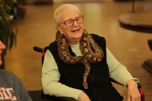 An elderly woman in a wheelchair smiles indoors with a snake draped around her neck, enjoying an Exotic Animal Safari experience.