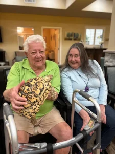 Two older adults sit together indoors, smiling at the camera as one holds a patterned tortoise, enjoying a mini Exotic Animal Safari experience. A walker is positioned in front of them.