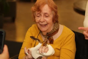 An elderly woman in a yellow outfit smiles while holding a white and brown snake during an Exotic Animal Safari, as others nearby observe and take photos.