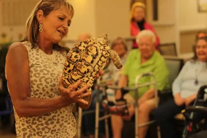 A woman holds a large patterned tortoise during an Exotic Animal Safari presentation, captivating an audience of seniors seated in a community room.
