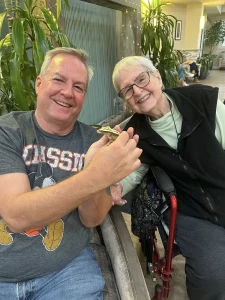 A man and an older woman smile as the man holds an exotic animal—a small lizard. The woman is seated with a red walker beside her. They are indoors, surrounded by green plants, creating a touch of safari adventure.
