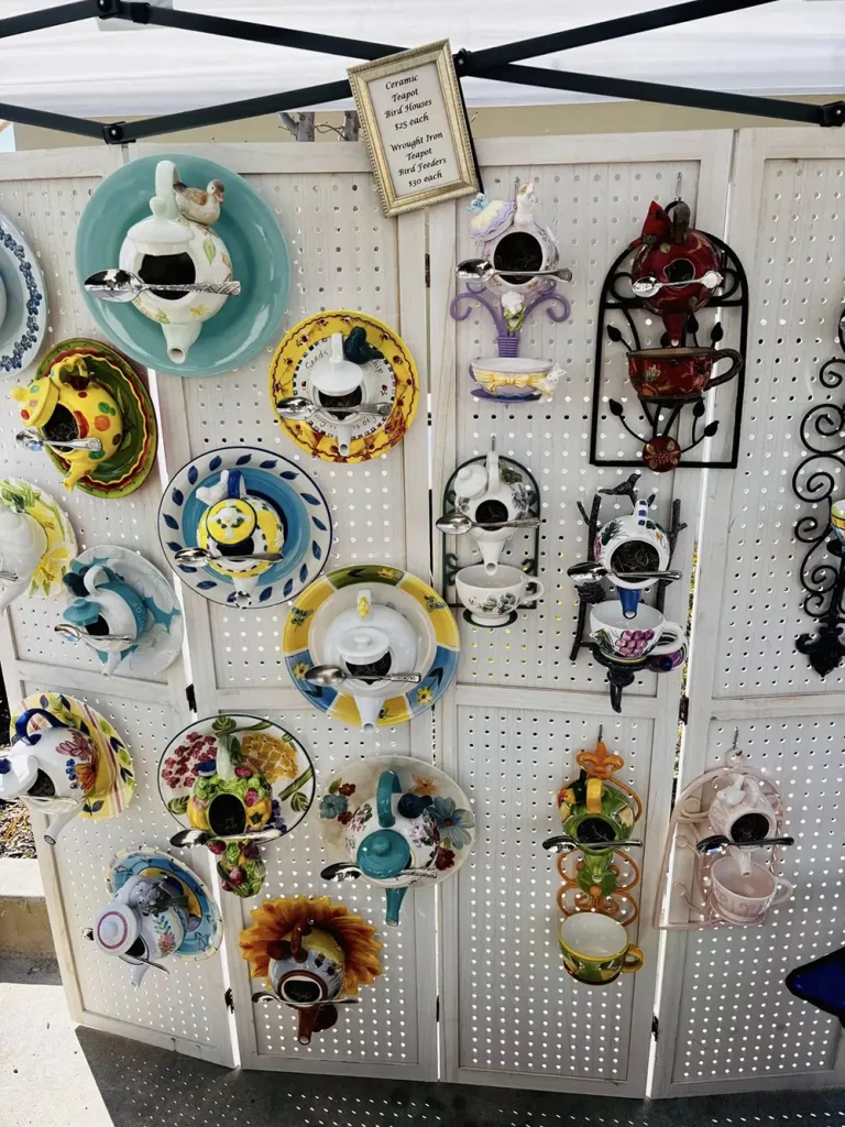 Colorful ceramic teapots and cups are mounted on a white pegboard display at the Spring Market, with a small sign above providing information about the items for sale.