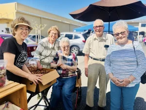Five older adults stand and smile together outdoors at the Spring Market, with a box of produce and an umbrella visible on a sunny day.