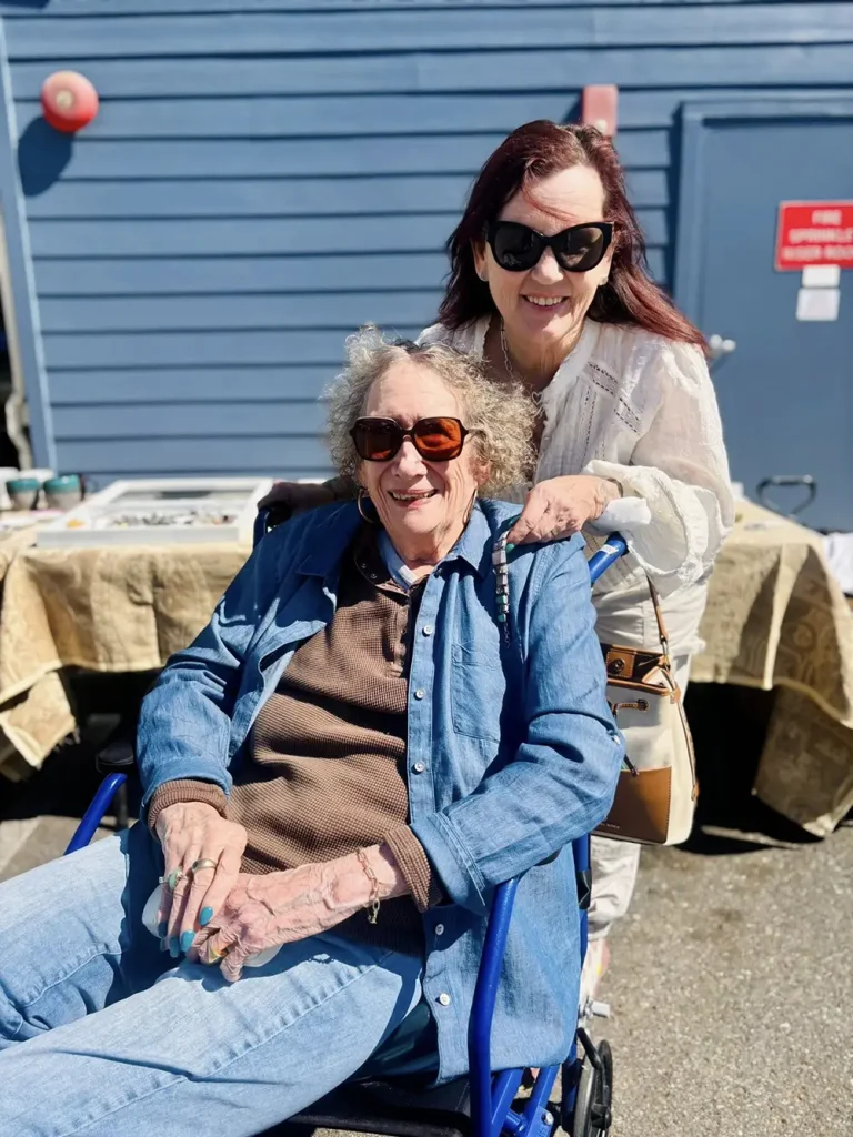 Two women smiling outdoors at the Spring Market; one is seated in a wheelchair wearing sunglasses and a denim outfit, while the other stands behind her, also sporting sunglasses.