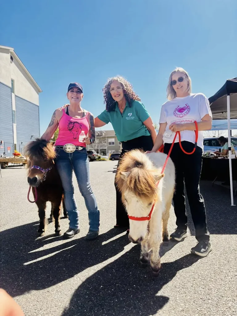 Three women stand outdoors with two miniature horses on leashes next to a building and market tents, enjoying the Spring Market under a clear blue sky.