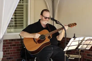 At a Spring Market Vendor Faire, a man wearing sunglasses plays an acoustic guitar and sings into a microphone while seated outdoors, with sheet music and an amplifier nearby.