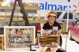 A woman stands behind a display of Ellie Marie's Gourmet Foods at an outdoor Vendor Faire, with packaged goods and a company sign visible. Walmart signage and parked cars are in the background.