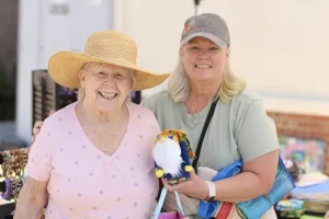 Two women stand outdoors at the Spring Market, smiling at the camera. One wears a sun hat; the other holds a small, bearded gnome figurine. Tables with items are visible in the background of this lively vendor faire.