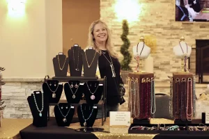 A woman stands smiling behind a display table filled with various necklaces and jewelry at an indoor Spring Market or Vendor Faire.