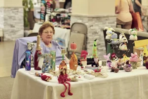 An older woman sits behind a table displaying various handmade crafts and decorations at the bustling Vendor Faire Spring Market.