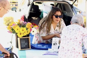 At a Spring Market, a woman wearing sunglasses chats with two older adults at an outdoor table adorned with flowers, brochures, and a sign reading, "Not all heroes wear capes. Some wear scrubs.