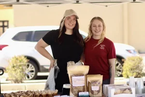Two women stand behind a market stall at the Spring Market, displaying packaged food products and baked goods outdoors, with a car and plants in the background.