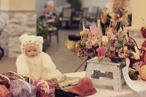 A doll in a white fuzzy outfit sits next to fabric bags and a display of small, colorful, handmade hanging dolls at the Spring Market Vendor Faire.