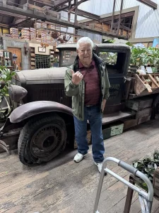 An older man stands indoors at High Hands Nursery next to a vintage pickup truck filled with plants and crates, pointing with his right hand and smiling at the camera, capturing a cheerful nursery outing.