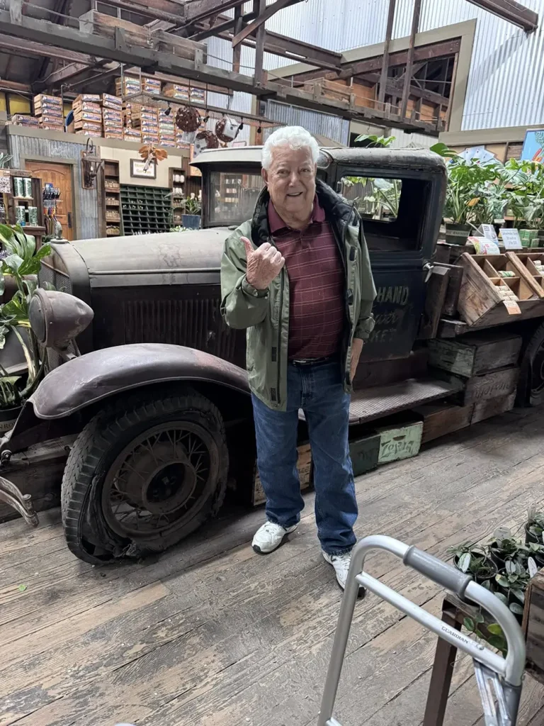 An older man stands indoors at High Hands Nursery next to a vintage pickup truck filled with plants and crates, pointing with his right hand and smiling at the camera, capturing a cheerful nursery outing.
