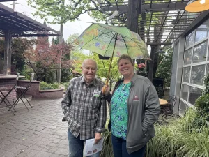 Two people stand outdoors on a stone path at High Hands Nursery; one person holds an umbrella over them both. Trees, plants, and outdoor seating from the nursery outing are visible in the background.