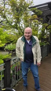 An older man in a beige jacket and jeans stands on a wooden path during a High Hands Nursery outing, surrounded by green trees and colorful flowers in the garden.