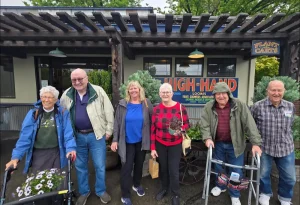Six older adults, some using walkers, stand smiling in front of High-Hand and Plant Café during a nursery outing to High Hands Nursery on a cloudy day.