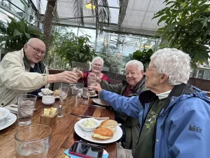 Four older adults sit around a wooden table with food and drinks, smiling and raising glasses in a toast during a nursery outing inside High Hands Nursery’s greenhouse-like setting surrounded by lush plants.