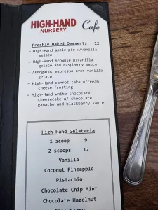 A dessert menu from High-Hand Nursery Cafe lists baked desserts and gelato flavors, with prices for single and double scoops—perfect treats for a High Hands Nursery outing. A fork is visible on the table beside the menu.
