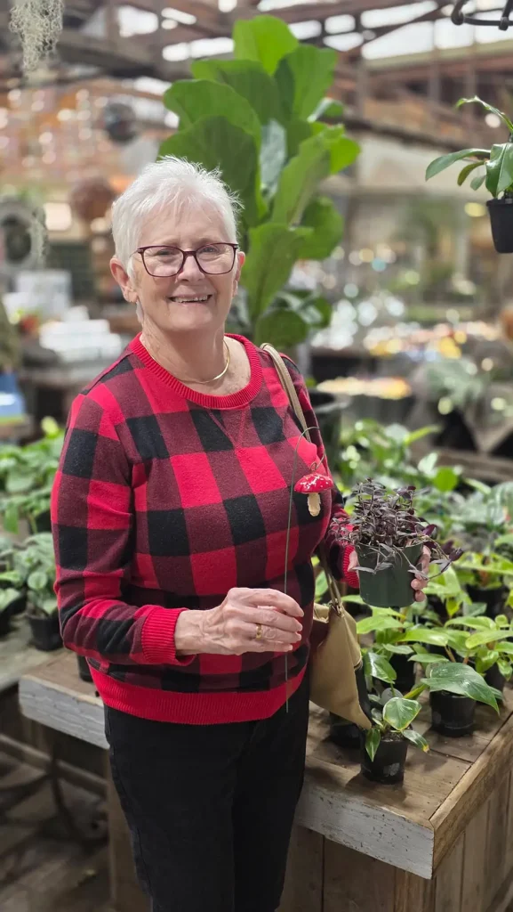 An older woman wearing glasses and a red plaid sweater stands in a greenhouse at High Hands Nursery, smiling and holding a small potted plant among various green plants during a pleasant nursery outing.