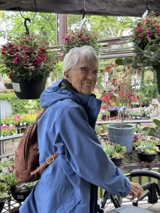 An older woman wearing a blue jacket and brown backpack stands among hanging and potted plants at High Hands Nursery, smiling at the camera during her nursery outing.