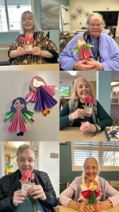 Elderly people in a care facility hold up handmade paper crafts, including colorful paper dolls and bouquets, celebrating Women empowerment and smiling for the camera on International Women’s Day.