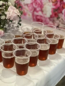 Several clear plastic cups filled with a brown beverage are arranged on a white tablecloth, with pink and white flowers in the background, creating a vibrant setting to celebrate International Women’s Day and support women empowerment.