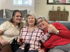 Three women sit closely together on a couch, smiling at the camera; one holds a drink, another is holding hands with her—capturing a joyful moment of women empowerment and friendship.