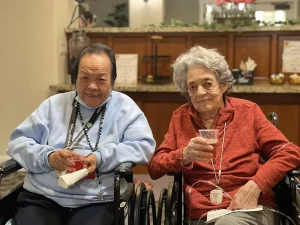 Two elderly women sit side by side in wheelchairs, each holding a drink and wearing lanyards, in a warmly lit indoor setting—celebrating International Women’s Day and their lifelong dedication to women’s rights.
