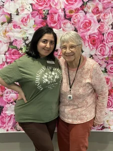 Two women stand side by side, smiling in front of a vibrant flower-patterned backdrop—a joyful moment celebrating women empowerment. One wears a green shirt and the other a pink top with a medical alert necklace.