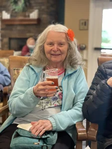 Elderly woman with long gray hair and an orange flower clip sits indoors, smiling and holding a drink in a clear plastic cup, embodying women empowerment in celebration of International Women’s Day.