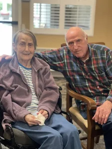 An older woman in a wheelchair sits beside an older man in a plaid shirt; both are looking at the camera indoors, reflecting the spirit of International Women’s Day and the importance of gender equality across generations.
