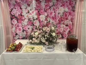 A table set for International Women’s Day features tea sandwiches, fruit skewers, plastic cups, a dispenser of iced tea, and a floral centerpiece before a pink rose backdrop.