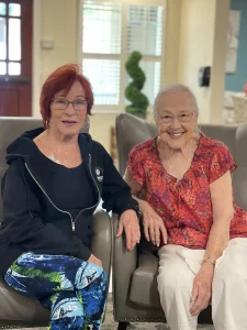 Two older women sit side by side indoors, smiling at the camera. Their warmth reflects women empowerment—one in a black jacket and patterned pants, the other in a red patterned blouse and white pants—celebrating strength and unity.