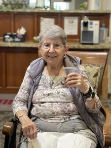 An older woman with short gray hair and glasses sits in a chair indoors, holding a cup and smiling at the camera, embodying women empowerment and the spirit celebrated on International Women’s Day.