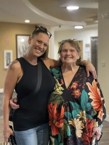Two women standing indoors, smiling at the camera. One has her arm around the other; both are wearing casual clothing, with the older woman in a floral patterned top—capturing a moment of women empowerment and friendship.